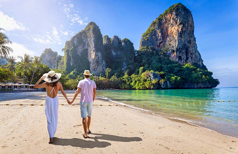 A happy couple on holiday walks along the empty Railay Beach in Krabi, Thailand, with emerald sea and lush rain forest. 